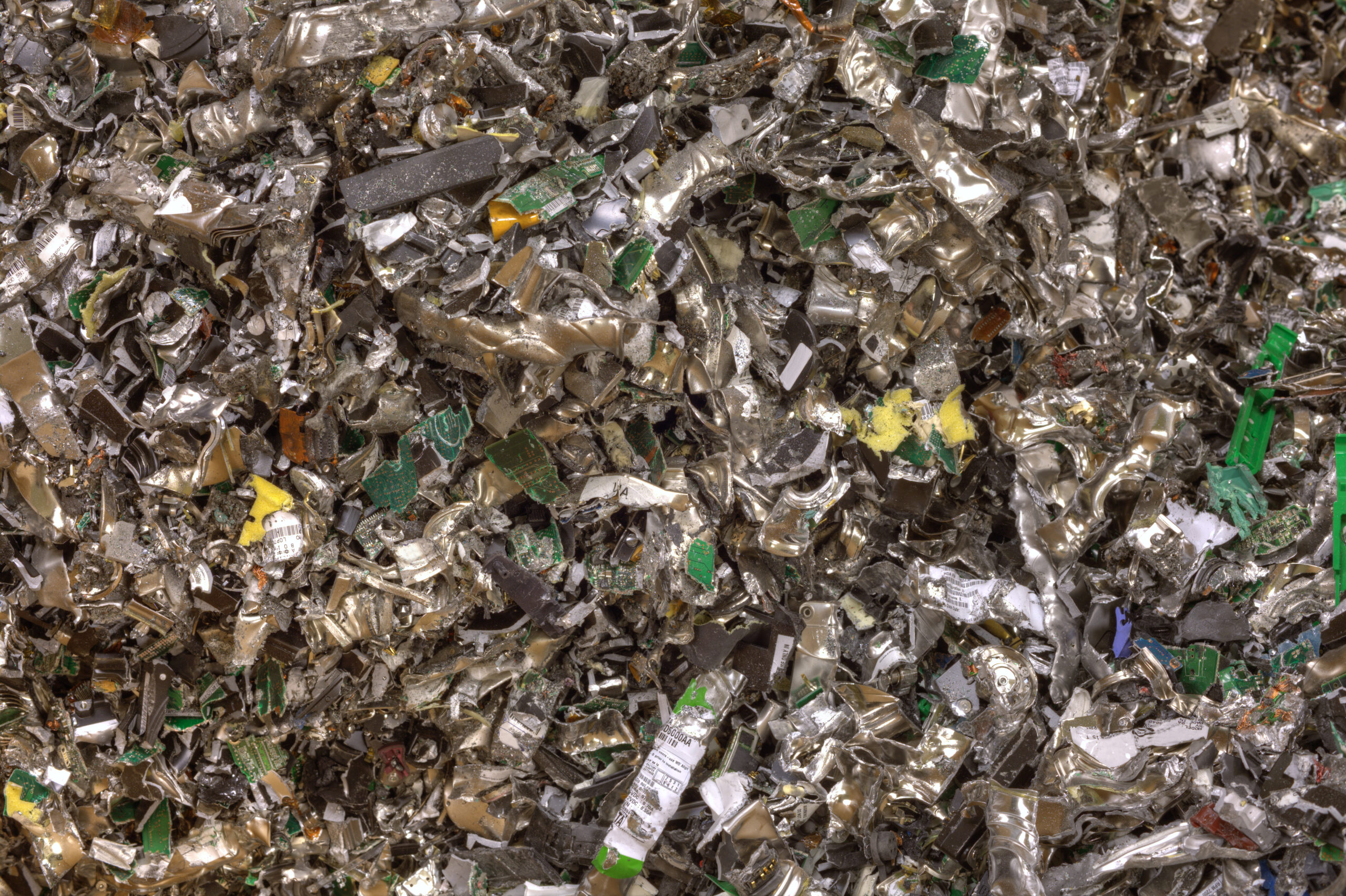 Pile of shredded electronic waste showing fragments of metal, green circuit boards, and plastic from recycled devices